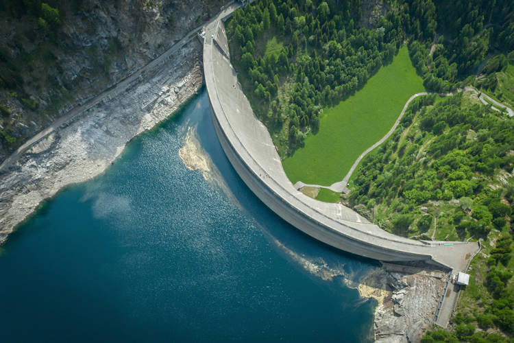 Green landscape surrounds the dam and water, country road visible below
