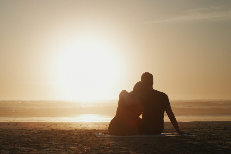 Couple, sunset and beach while on a romantic nature date watching ocean view on a picnic during their vacation. Love, travel and summer holidays with lovers in silhouette from behind for a honeymoon