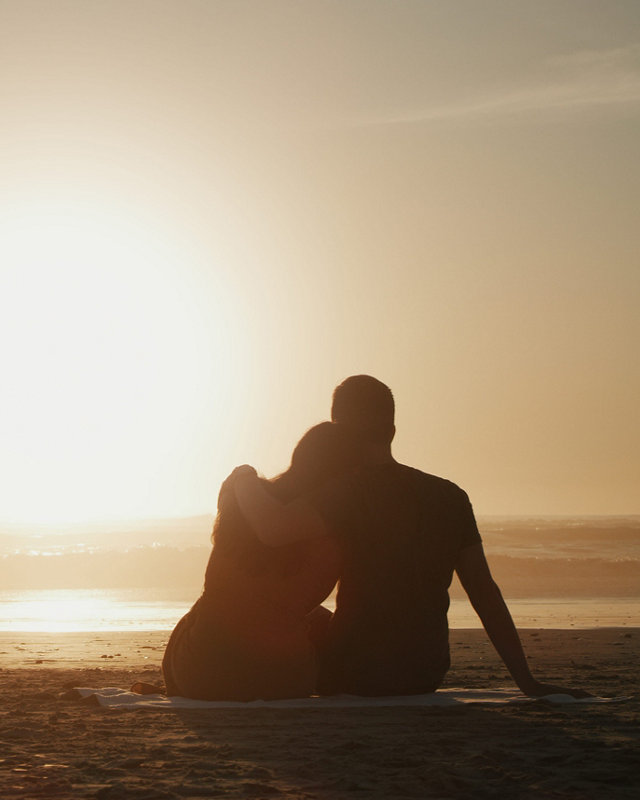 Couple, sunset and beach while on a romantic nature date watching ocean view on a picnic during their vacation. Love, travel and summer holidays with lovers in silhouette from behind for a honeymoon