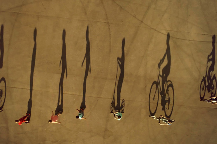 A group of preadolescents children dressed in a summer casual clothes are riding the eco alternative city transport (bikes, scooter, roller skates and self balancing scooter) at the walk on city square. Aerial top view, drone shot, outdoors in a summer at a sunset with large shadows