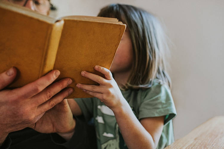 A little girl looks enthusiast and interested as her father holds a yellow book at reads it to her. She looks into the book as he reads.