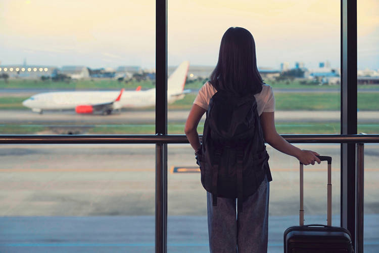Travel tourist standing with luggage watching sunset at airport window. Woman looking at lounge looking at airplanes while waiting at boarding gate before departure. Travel lifestyle. Transport and travel concept