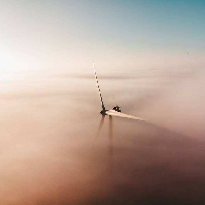 Wind turbine in foggy landscape, casting shadow. Soft light creates serene atmosphere. Sky transitions from blue to peach.
