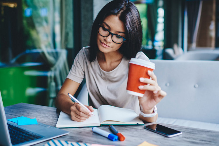 Concentrated female student writing in notebook while learning with cardboard coffee cup in cafe, pensive woman freelancer noting information for planning project doing remote job via laptop computer; Shutterstock ID 1389068438; Nom de Projet: Pictet AM