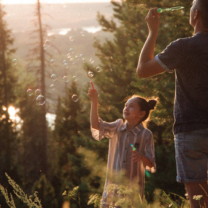 Two people enjoy blowing bubbles in a sunlit forest, surrounded by trees and a distant view of a lake.
